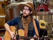 A man wearing a khaki scarf and large hat plays guitar and sings while sitting on a barstool inside a stage at a brewery.