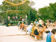 A band plays on stage outside to a crowd of people sitting on picnic tables.