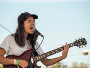 Young woman with a black baseball hat on plays guitar while singing into the microphone.