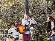 A bland plays on stage with two male guitarists, one male drummer, and a female guitarist who is singing into the microphone.