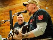 Two men play guitar while sitting on stools inside a brewery.