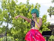 A girl with a bright blue wig on and neon pink skirt sings into the microphone while pointing at the crowd. 
